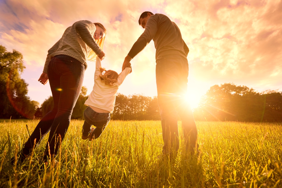 A mother and father swing their child between them by their arms. The sun sets behind them.